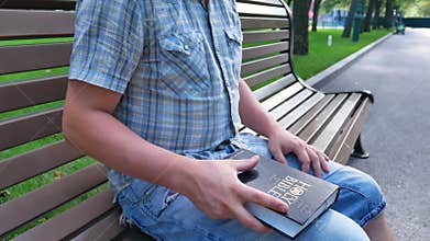 Man is holding Holy Bible in his hand sitting on a bench in a park in the summer on a sunny day