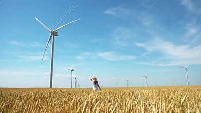 Beautiful girl walking on yellow field of wheat with windmills for electric power production