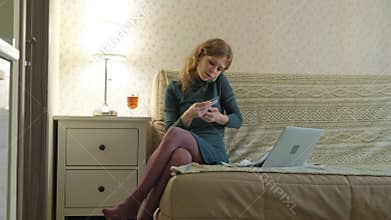 A young woman in a green dress sits on a sofa with a laptop, uses a telephone and a plastic card, works, studies