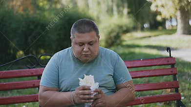 A fat man eats and looks at his harmful and tasty fast food on a bench