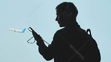 A young guy turns on music on a smartphone and puts on headphones against the background of a plane taking off.
