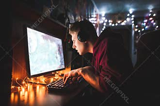 Concentrated teenager sits at a computer table in the cozy room, looks at the monitor and plays video games