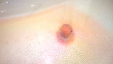 Small colorful bath bomb lies on the bottom in the tub and slowly dissolves. On the surface of the water traces of aromatic oil.