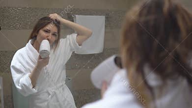 Funny charming girl in white robe took bath and sings in front of mirror with hairdryer.