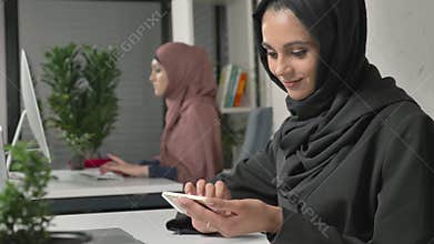 Young beautiful girl in black hijab sits in office and uses smartphone. Girl in black hijab in the background. Arab