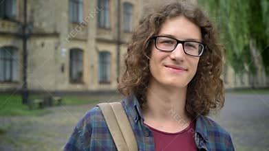 Young handsome happy student in glasses and with backpack, standing near on street near college campus, smiling