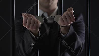 Man in suit showing hands in handcuffs behind bars, bribery, financial fraud
