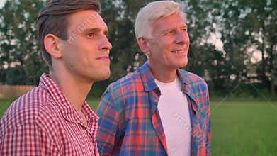 Old father hugging adult son and looking forward, standing on wheat or rye field, forest in background