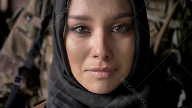 Close portrait of young muslim woman in hijab crying and looking at camera, armed soldier with weapon standing behind