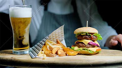 Chef is serving set of tasty burger, french fries and beer on the wooden tray on the black background