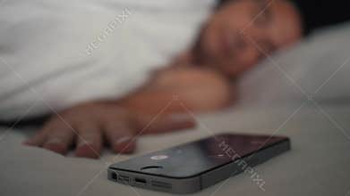 Young man sleeping on the bed with an alarm clock on the mobile phone display. Modern technologies
