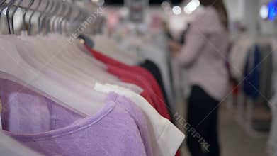 Clothes shop, fashionable new clothing hanging on hangers and shopper woman in unfocused background chooses purchases in