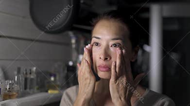 Asian middle aged woman applying face cream in the kitchen