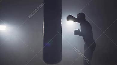 Boxer hits punching bag in dark gym. Young man training indoors