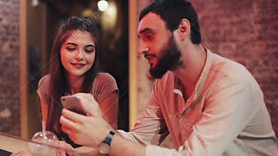 Happy young couple with smartphone talking and drinking beverage sitting at the bar. Friends using cell phone smartphone