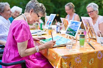 Stylish senior lady painting in art class with friends from her care home for the aged copying a painting with water colors.
