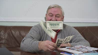 Happy old man throwing away the dollars sitting at the wooden table close up. Positive rich man demonstrates his money