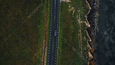 Aerial top view tracking shot of black car moving along epic highway road along old autumn eroded sea bay coastline.