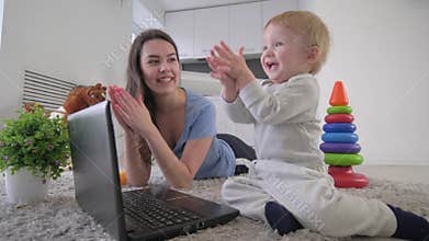 Child development, curious merry infant boy with young mom push buttons laptop computer and clap hands lying on floor