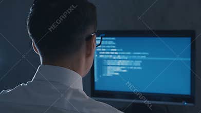 Back view of programmer professional in glasses programming code on computer monitor at night office