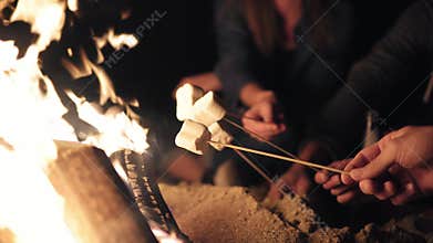 Closeup view of marshmallow on sticks being fryed by the bonfire. Group of people sitting by the fire late at night