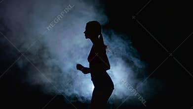 Athletic fitness girl running on a black background illuminated by the spotlight in the smoke. Silhouette. Slow motion