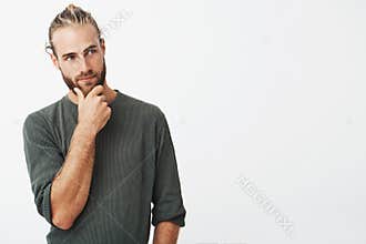 Attractive swedish guy with stylish hair and beard in grey shirt holding his chin and thoughtfully looking aside