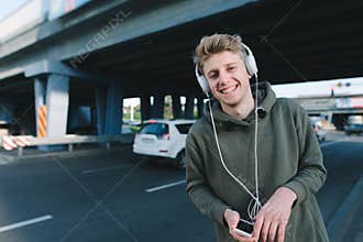 Street portrait of a happy young man who smiles, and listens to a musician in headphones in the background of urban architecture.