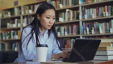 Young asian student girl sitting at table with pile of books in university library working on laptop computer, drinking