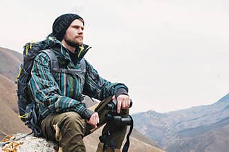 Tired bearded hipster with binoculars in his hands sits on a stone among the mountains and looks out into the distance