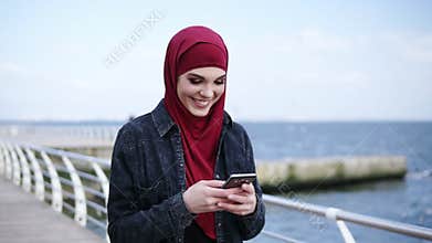 Attractive young girl with hijab on her head is smiling while texting to someone and scrolling something on her