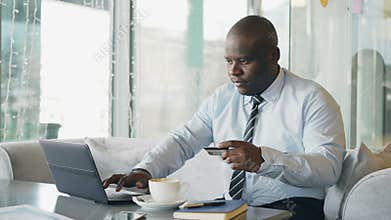Smiling African American businessman in formal clothes paying online bill using his credit card and laptop in
