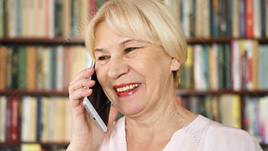 Modern senior woman at home talking on cellphone in library. Bookcase bookshelves in background
