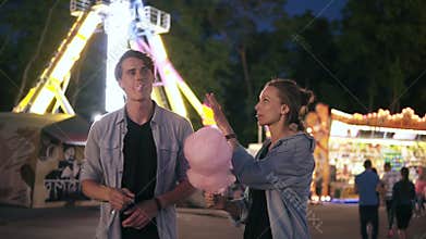 Young hipster couple are having time together in the amusement park at night. Feeding each other with pink cotton candy