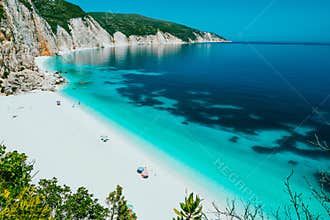 Sunny Fteri beach lagoon with rocky coastline, Kefalonia, Greece. Tourists under umbrella chill relax near clear blue