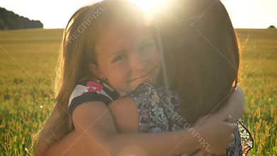 Happy beautiful little daughter hugging mother and smiling at camera, wheat or rye field during sunset in background