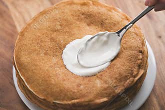 Adiing sour cream to stack of fresh hot crepes or blinis on wood table