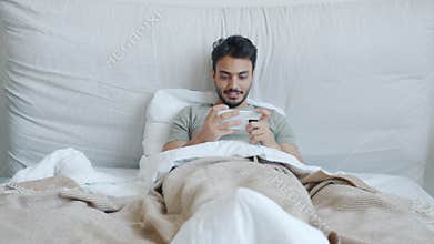Attractive Arab man playing video game with smartphone lying in bed alone