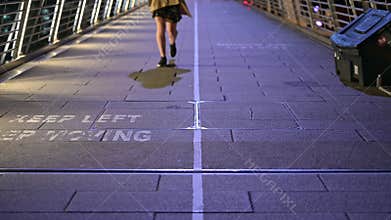 A woman`s legs walking the wrong way across a KEEP LEFT sign painted on Jubilee Foot Bridge at night