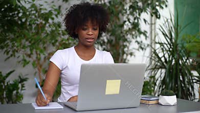 Professional mixed race young female freelancer video conference calling on laptop computer, making notes, talking by