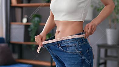 Closeup slim unrecognizable female abs wearing big jeans measuring waist size with measurement tape