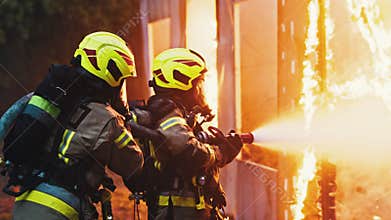 Close up, two firefighters extinguish fire, spraying house with hose at night.