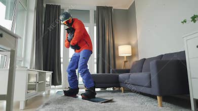 Fun video. Man dressed as a snowboarder rides a snowboard on a carpet in a cozy room. He holds a black cat in his arms