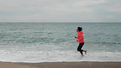 Athletic woman is jogging along sandy beach against stormy ocean with big waves. Happy carefree female is running barefoot on wave