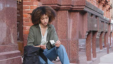Frustrated upset young African American woman with backpack sitting on stairs outdoors holding dollar bills. Ethnic