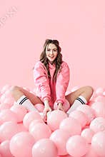 Pretty smiling woman sitting on floor with lots of pink balloons on pink background