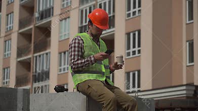 Construction worker using smartphone on coffee break on construction site. Builder resting at project residential area