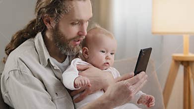Adult caucasian father bearded daddy with little daughter son infant newborn sitting at home sofa looking at mobile