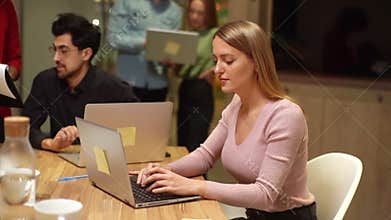 Tracking shot of focused young business woman sitting at office desk and working on laptop while multiethnic team of
