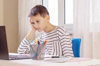 Sad tired boy sitting at table with books and laptop computer at home. Learning difficulties, online education concept